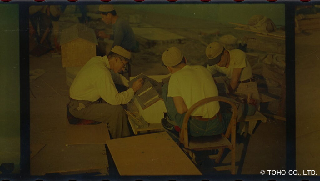 帽子を被った複数のスタッフたちが低い椅子に座り、木製の日本家屋と思われるミニチュア模型の屋根瓦や細部に筆で丁寧に塗装や修正を施している様子を捉えた当時の特撮映画製作の写真
