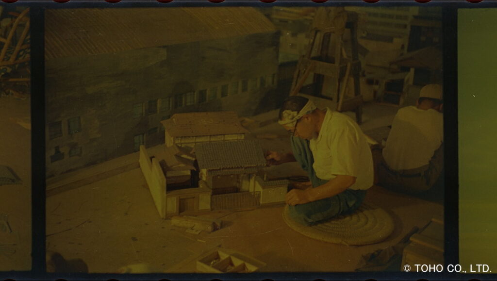 薄暗い照明の作業場において、鉢巻を締めた眼鏡のスタッフが円座に座り、瓦屋根や白壁が再現された日本家屋の精巧なミニチュア模型に対し、筆を使って細部の塗装や修正作業を行っている当時の写真