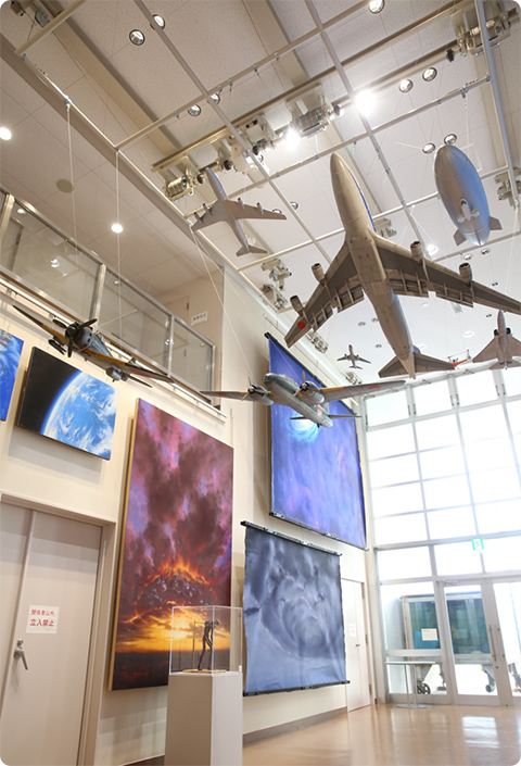 特撮用の戦闘機や飛行船のミニチュア模型が天井から吊るされた展示室。壁面には雲海や爆発シーンを描いた大きな背景画が並び、特撮美術の世界を再現している。