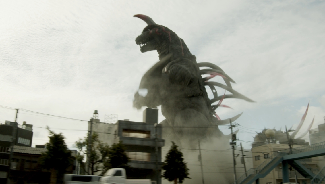 住宅街のビルや電柱を見下ろすほど巨大な、一本角と背中に複数の鋭い突起を持つ怪獣が、土煙を上げながら街中を闊歩している、特撮映画のワンシーンのような迫力ある写真