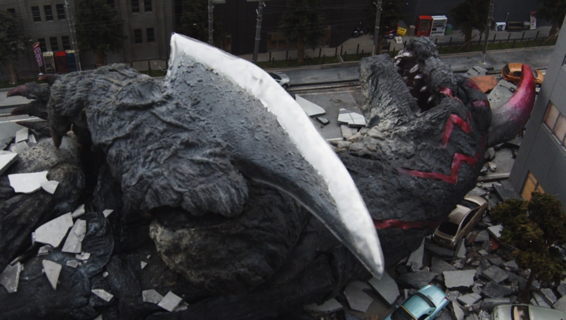 瓦礫が散乱する街の中に、鋭い刃物のような前足と赤い一本角を持つ巨大な怪獣が倒れ込み、破壊された道路や建物、停車した車を見下ろすようなアングルでその巨大さを強調して捉えた、特撮映画のワンシーンのような臨場感溢れる写真