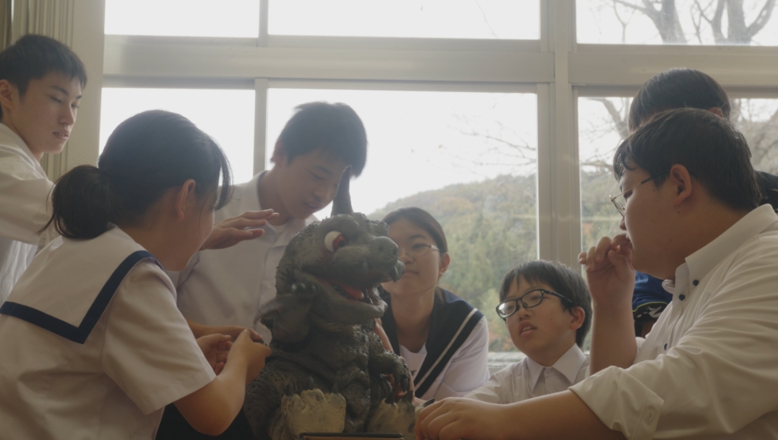 明るい窓際で、制服を着た数名の学生たちが机を囲み、中央に置かれた小さな怪獣のフィギュアを興味深そうに覗き込んだり、手を添えて調整したりしている様子を捉えた写真