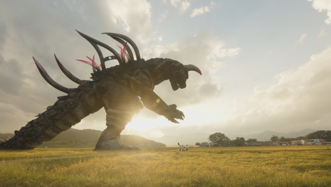 夕日に照らされた広大な黄金色の田んぼの中に、背中にいくつもの鋭い角を持つ巨大な怪獣が立ち、その足元には豆粒ほどに小さく見える数人の人物が向かい合っている写真