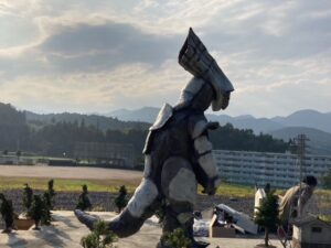 屋外の特撮セットで、特徴的な頭部を持つ巨大な怪獣の着ぐるみが、ミニチュアの樹木や建物の間を縫って静かに佇んでいる様子の写真