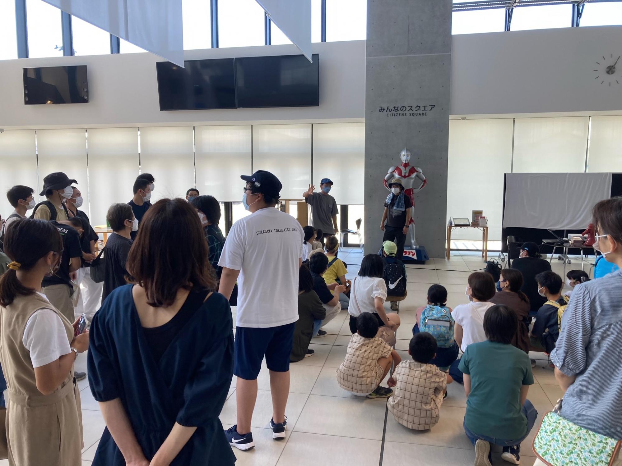 明るく開放的な「みんなのスクエア」という屋内施設で、フロアに集まった多くの人々が、マスクを着用しながら前方の説明を行う男性の話を熱心に聞いている写真