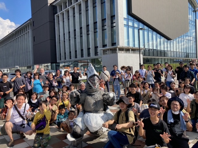 須賀川市役所を背景に、広場に集まった大勢の老若男女が怪獣の着ぐるみを囲んで笑顔でポーズを決めている集合写真