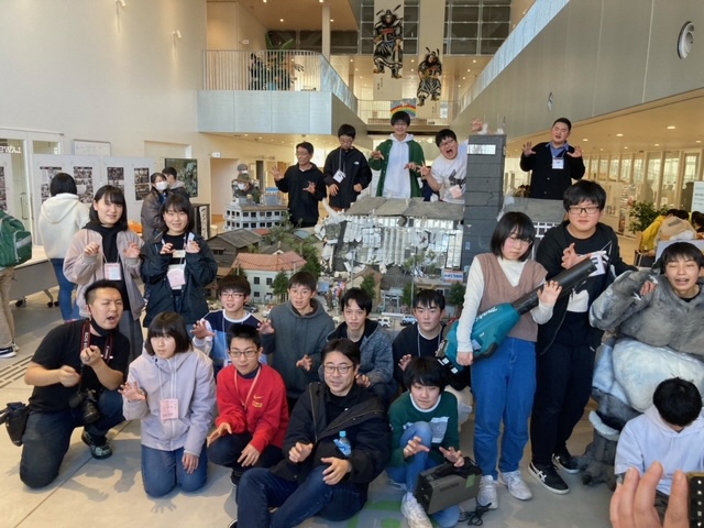 吹き抜けのある広々としたロビーで、精巧に作られたビルや住宅のミニチュアセットを囲み、多くの参加者たちが怪獣を模したポーズをとったりしている「すかがわ特撮塾 第1期生 制作映画上映会」の集合写真