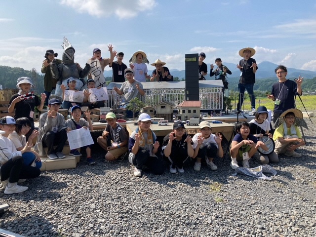 晴天の屋外でミニチュアの街並みを囲み、怪獣の着ぐるみや怪獣の設計図のイラストを手にした大勢の塾生たちと講師が、カメラに向かって爪を立てるようなポーズをして写した集合写真