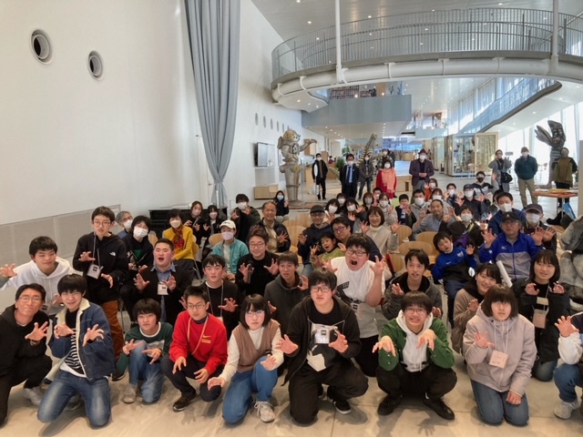 吹き抜けのあるホールで、大勢の若者や後列に立つ大人たちが、両手を爪のように立てるポーズをしている集合写真