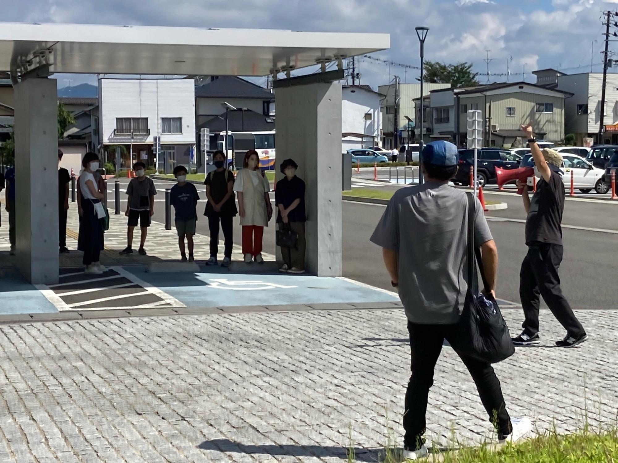 晴天の屋外にある石畳の広場で、赤いメガホンを持った男性が前方の屋根の下に並ぶ数人の男女に向かって合図を送っている様子を後方から捉えた写真