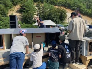 晴天の屋外で、数名の塾生が大きな灰色の建物模型の内部を覗き込んだりして、手を加えたりしながら確認をしている写真