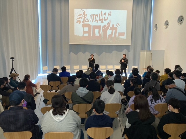 スクリーンに「魂の叫び ヨロイガー」という力強い筆文字のタイトルが投影され、その前で二人の男性がマイクを手に、椅子に座った多くの観客に向けて話をしている写真