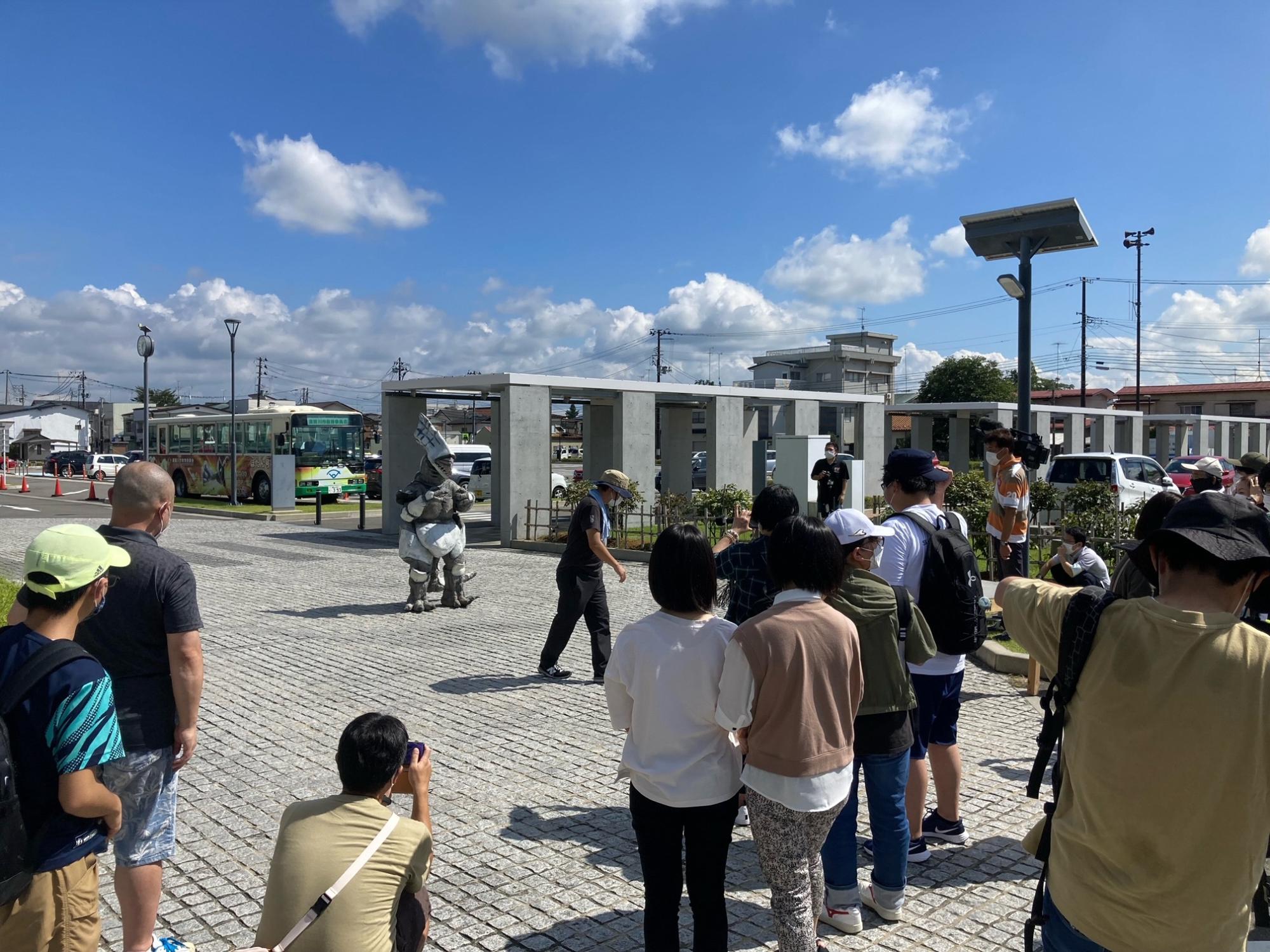 青空が広がる屋外の石畳の広場で、怪獣の着ぐるみが歩いており、周囲に集まった多くの人々がスマートフォンで撮影したり見守ったりしている様子の写真