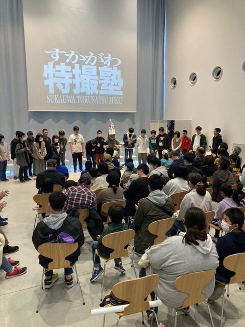 ホールに集まった「すかがわ特撮塾」の塾生たちが、正面のスクリーンに投影された塾のロゴを背景に一列に並び、手前の椅子に座った多くの観客に見守られながら、制作した作品の発表や修了の挨拶を行っている様子の写真