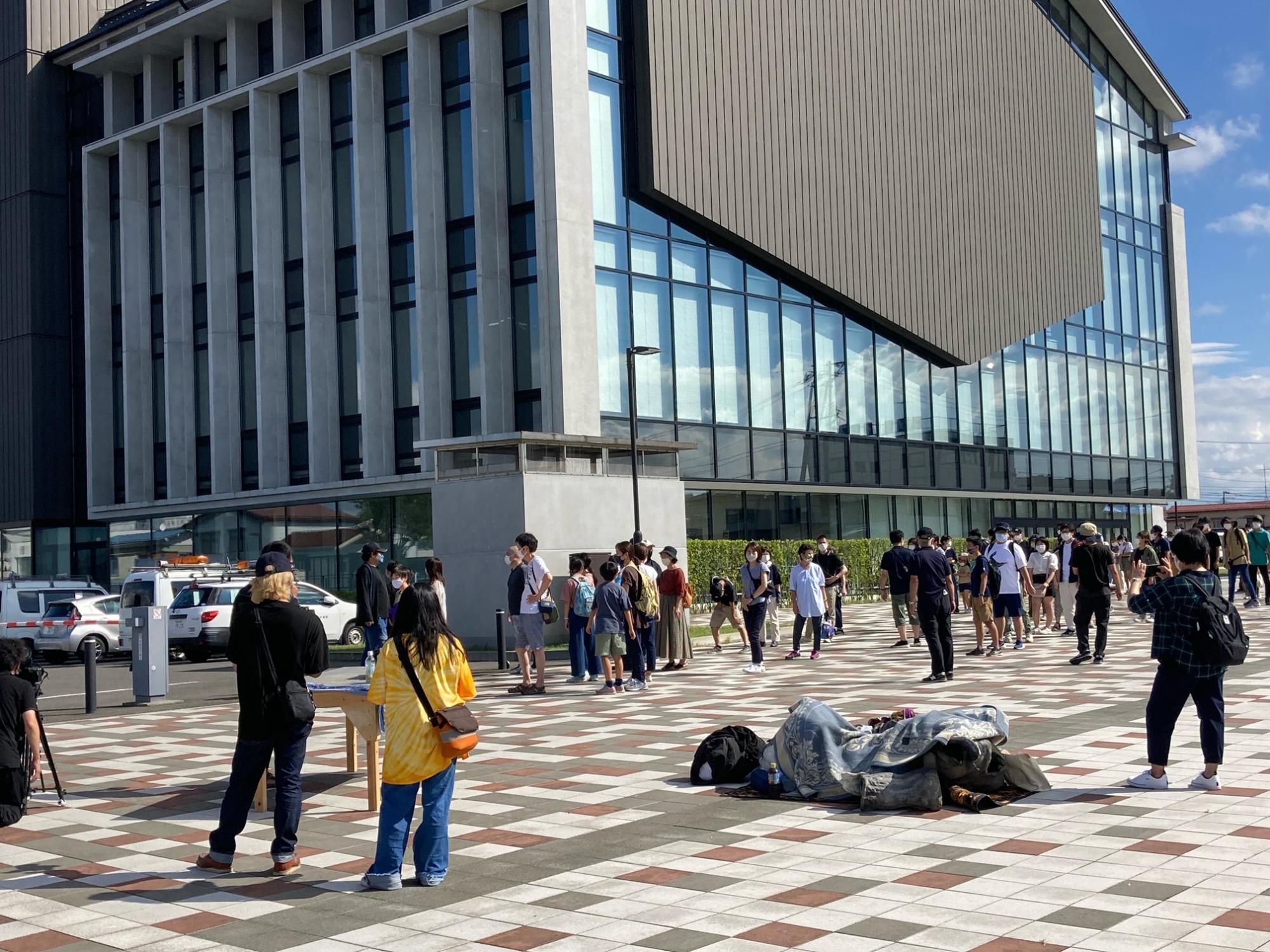 須賀川市役所を背景にした広々としたタイルの広場で、多くの人々が等間隔に並んだり移動したりしており、手前の地面には毛布のような荷物が置かれている写真