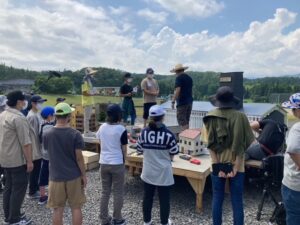 屋外にて、ミニチュアの街並みセットの周囲に多くの塾生が集まり、ミニチュアの街並みセットの建物の前で講師が説明しているのを3人の塾生が聞いている様子の写真