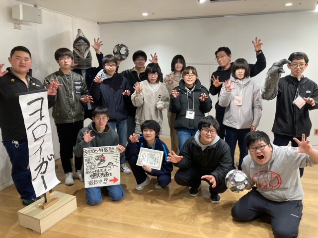 明るい室内で「すかがわ特撮塾 第一期生」に参加した参加者たちが、怪獣を模したポーズをとったり自作のイラスト看板や修了証を掲げたりしている集合写真
