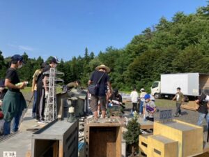 山林を背景にした屋外の広場にミニチュアの鉄塔や建物が置かれ、特撮の準備を進めている写真