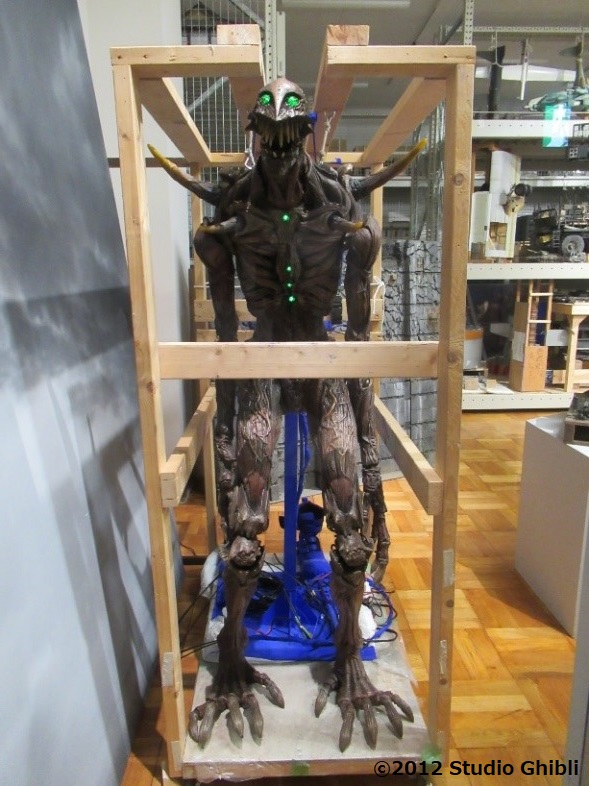 木製の骨組みの中に直立し、緑色に発光する目と胸部のランプ、そして全身に張り巡らされた筋肉のような質感や鋭い爪を持つ足先が特徴的な、特撮映画で使用される巨神兵の全身造形物を正面から捉えた写真