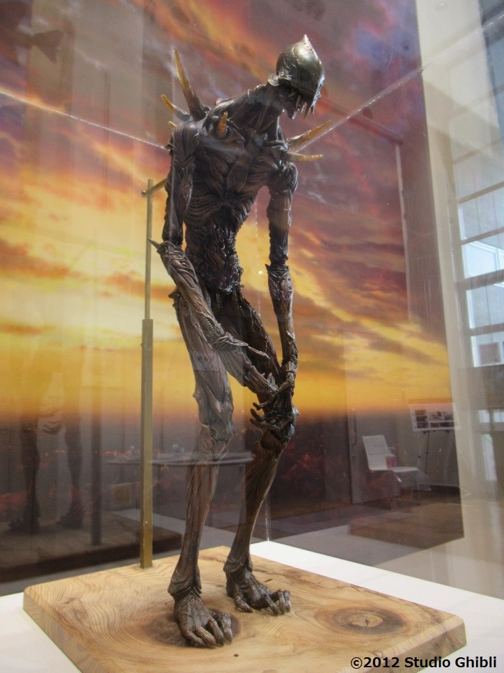 ガラスケースの中、木の板の上で少し猫背の姿勢で立っている背の高い巨神兵のパペットの写真