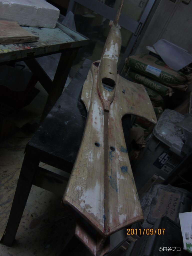 薄暗い倉庫のような作業台の上に置かれた、塗装が剥げ落ち木目が剥き出しになった大型戦闘機模型の写真