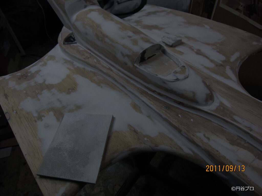 木材の表面を白いパテで広範囲に埋め立て、コックピット周辺の滑らかな曲線や機体の凹凸を整えている修復工程途中の写真