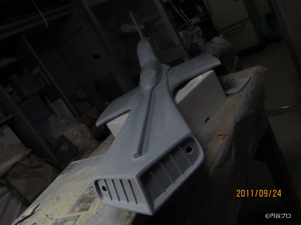 薄暗い作業室内で、グレーで下地塗装が施された大型戦闘機模型を前方から捉えており、機首部分の吸気口のようなスリット造形や滑らかに整えられた機体表面の質感が分かる写真