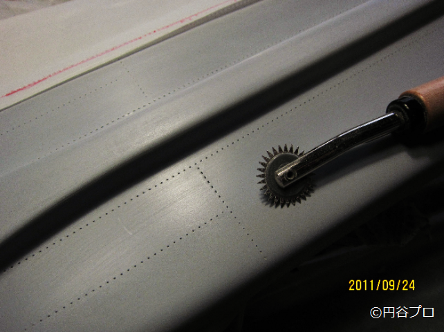 下地塗装が施された大型戦闘機模型のグレーの機体表面に、ステッチルレットを使って機体のリベットを表現している部分を至近距離から撮影した写真