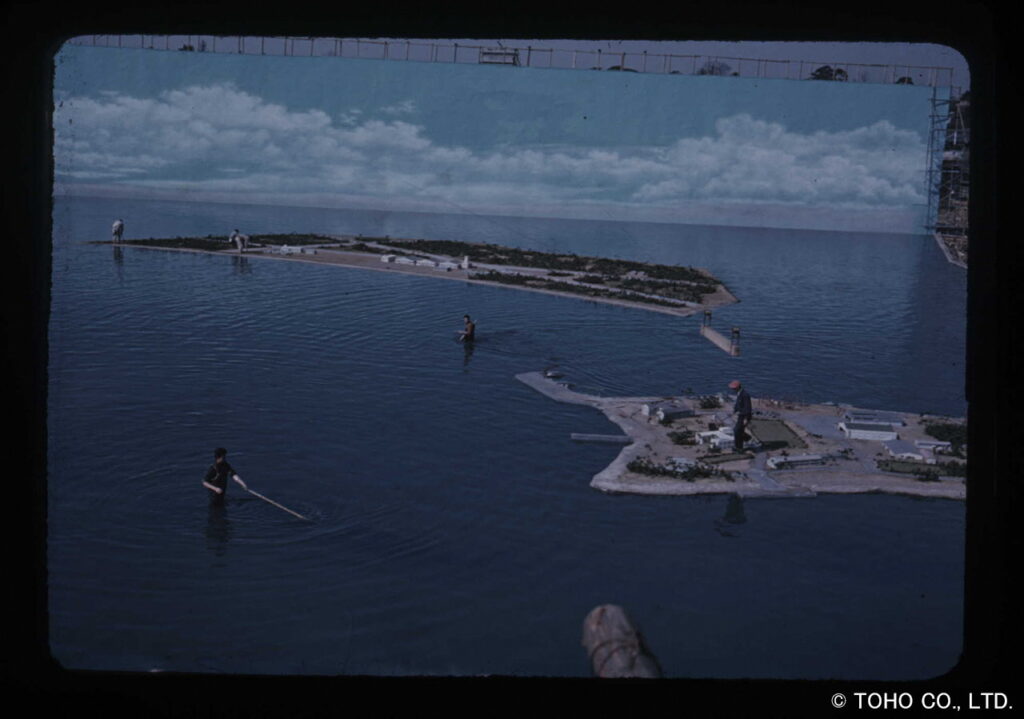 巨大なプールのような特撮用オープンセットで、水面に浮かぶミニチュアの島々や背景に設置された青空の描き割りを背景に、腰まで水に浸かったスタッフたちが撮影の準備や調整を行っている様子を捉えた、当時のセットでの製作現場を記録した写真