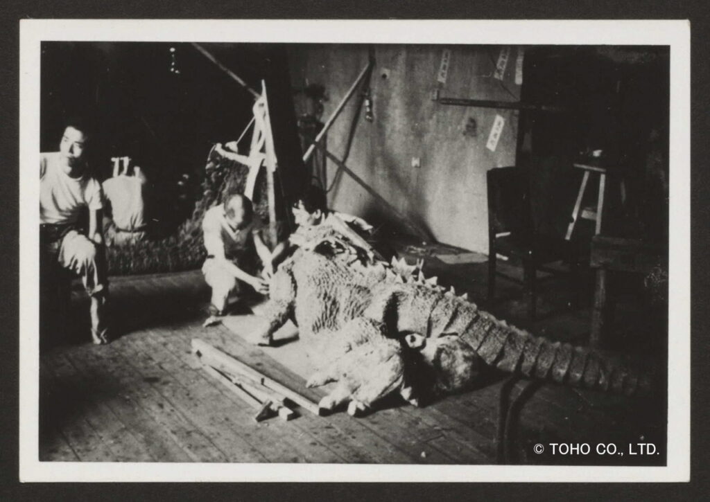 数名のスタッフがしゃがみ込んで、床に横たわった巨大なゴジラの着ぐるみの製作を行っている当時の制作現場の様子を記録したモノクロ写真