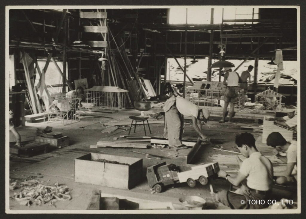 映画の特撮用ミニチュア制作現場にて、複数のスタッフが建物の骨組みや車両の模型を丁寧に組み立てており、木材や道具が散乱する工房内で緻密な手作業が進められている様子を捉えた、当時の風景を記録したモノクロの写真