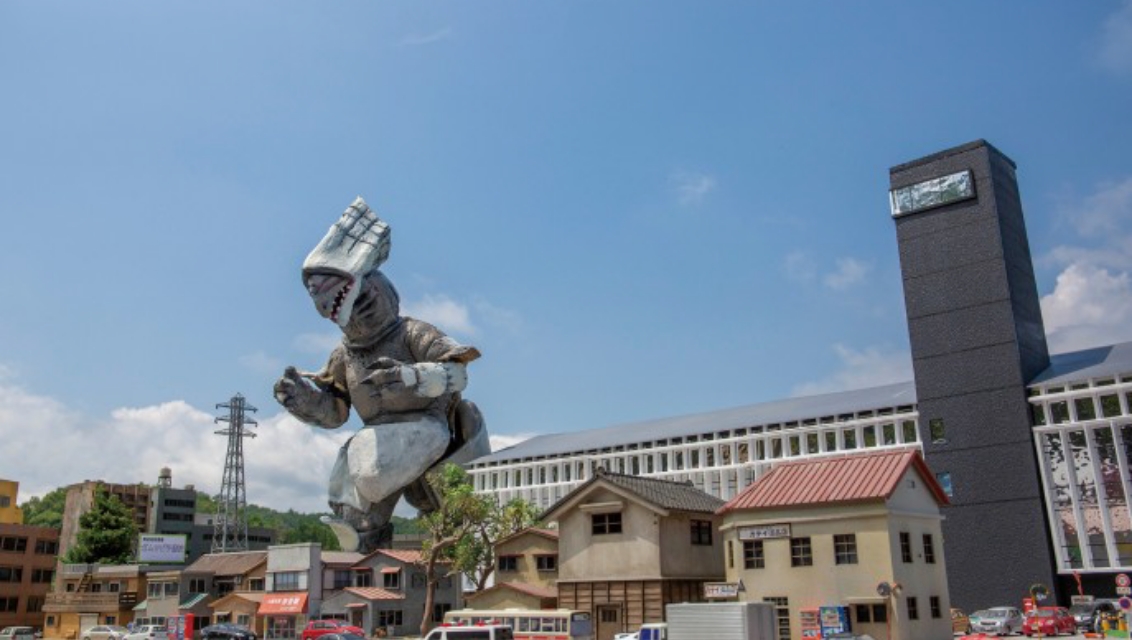 巨大な怪獣スーツが精巧なミニチュア都市の建物群を見下ろすように立ち迫力ある姿を見せる写真