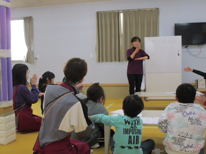 ホワイトボードの前に立つ女性講師が手本を見せ、それに向かい合って座る子供たちが真似をしながら一緒に手話を学んでいる出前講座の写真