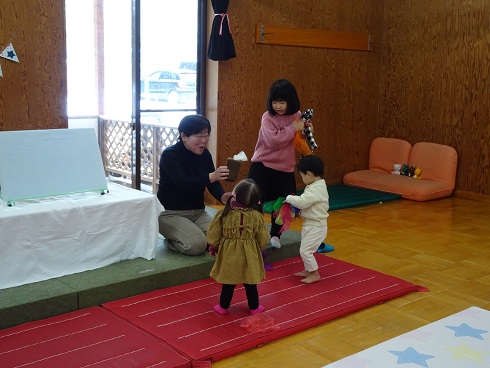 緑のマット上に座る人物が白い紙コップを持ち、前方には赤いマットの上で茶色の服を着た子どもと白い服を着た子どもが立ち、奥にはピンクの服を着た子どもがカラフルな布製アイテムを手にしている様子の写真