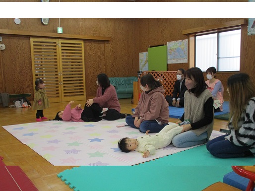 複数の人々が座って子どもと過ごしており、中央には白い服を着た赤ちゃんが仰向けになっていたり、奥には茶色の服を着た子どもが立っている様子の写真