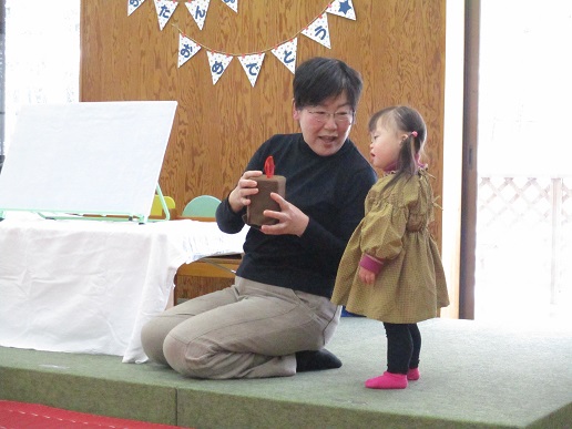 黒い服を着た人物が膝をつき、手にろうそくに見立てた茶色の箱型のアイテムを持ち、隣には茶色の服とピンクの靴下を身につけた子どもが立っている写真