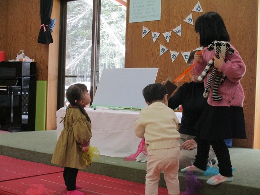 赤いマットの上に茶色の服を着た子どもと白い服を着た子どもが立ち、カラフルな布を手にして遊んでおり、隣にはピンクの服を着た子どもが縞模様のぬいぐるみを抱えている様子の写真