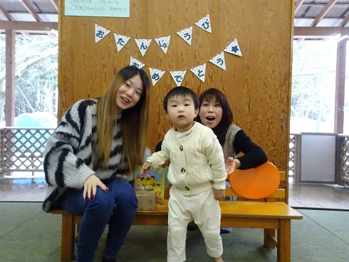 壁を背景に「おたんじょうびおめでとう」と書かれたガーランドが飾られ、緑色のマット上に木製ベンチが置かれ、白い服を着た子どもがベンチの前に立っている様子の写真