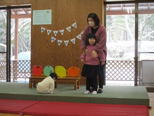 緑のマット上に赤・黄・オレンジの丸い紙が貼られたベンチが置かれ、その前でピンクの服を着た子どもが大人と一緒に立ちおわりのことばを言っている様子の写真