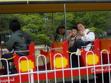 屋外で赤と黄色の屋根付き車両に複数の人が座り、車両の側面には白い文字と黄色の装飾があり、遊園地の豆汽車の写真