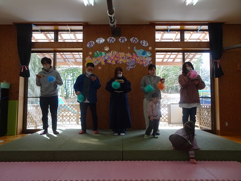 室内のステージ上で6人が並び、色とりどりのポンポンを手に持って立っており、背景には「おめでとう」の文字と花や鳥の装飾があり、前方には床に座る子どもが見える写真