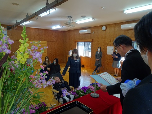 中央で車いすに乗っている子どもと濃紺の服を着た人物が立ち、手前では保育証書を持つ人物がいる様子の写真