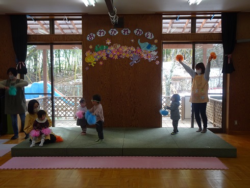 ステージ上で複数の子どもと大人が色とりどりのポンポンを手に持ちお遊戯をしている様子の写真