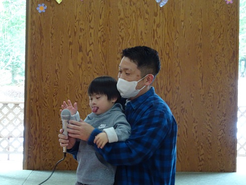 チェック柄のシャツを着た人物が幼い子どもを抱き、子どもが両手でマイクを持ってい「はじめのことば」を話している様子の写真