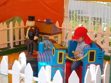 白い柵で囲まれた屋外の遊具エリアで、赤い帽子をかぶった子どもがトーマスの乗り物に座り、前方には黒い服を着たトップハム・ハット卿の人形と小さな家の模型が置かれている写真