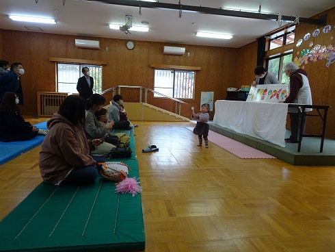 白い布を掛けたテーブルで絵本読み聞かせをしている人達が立ち、中央には小さな子どもが歩いている様子の写真