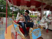 屋外のメリーゴーランドで、赤い座席に親子二人が乗り、周囲には白い馬の装飾が並び、座席の側面には青と金色の模様が描かれているのが見える写真