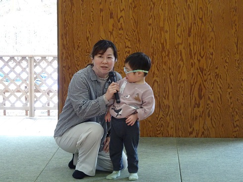 チェック柄の服を着た人物が膝をつき、隣に立つ幼い子どもと一緒にマイクを持って「おわりのことば」を話している様子の写真