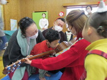 室内で赤い服の人物がギターを抱え、複数の人物が集まり楽器に手を添えている様子が見える写真