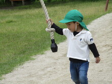 緑色の帽子をかぶり、白い長袖シャツと青いジーンズを着た子どもが、砂地の遊び場で太いロープの先端を片手で持ち、もう一方の手を横に広げて歩いている様子が写っている写真